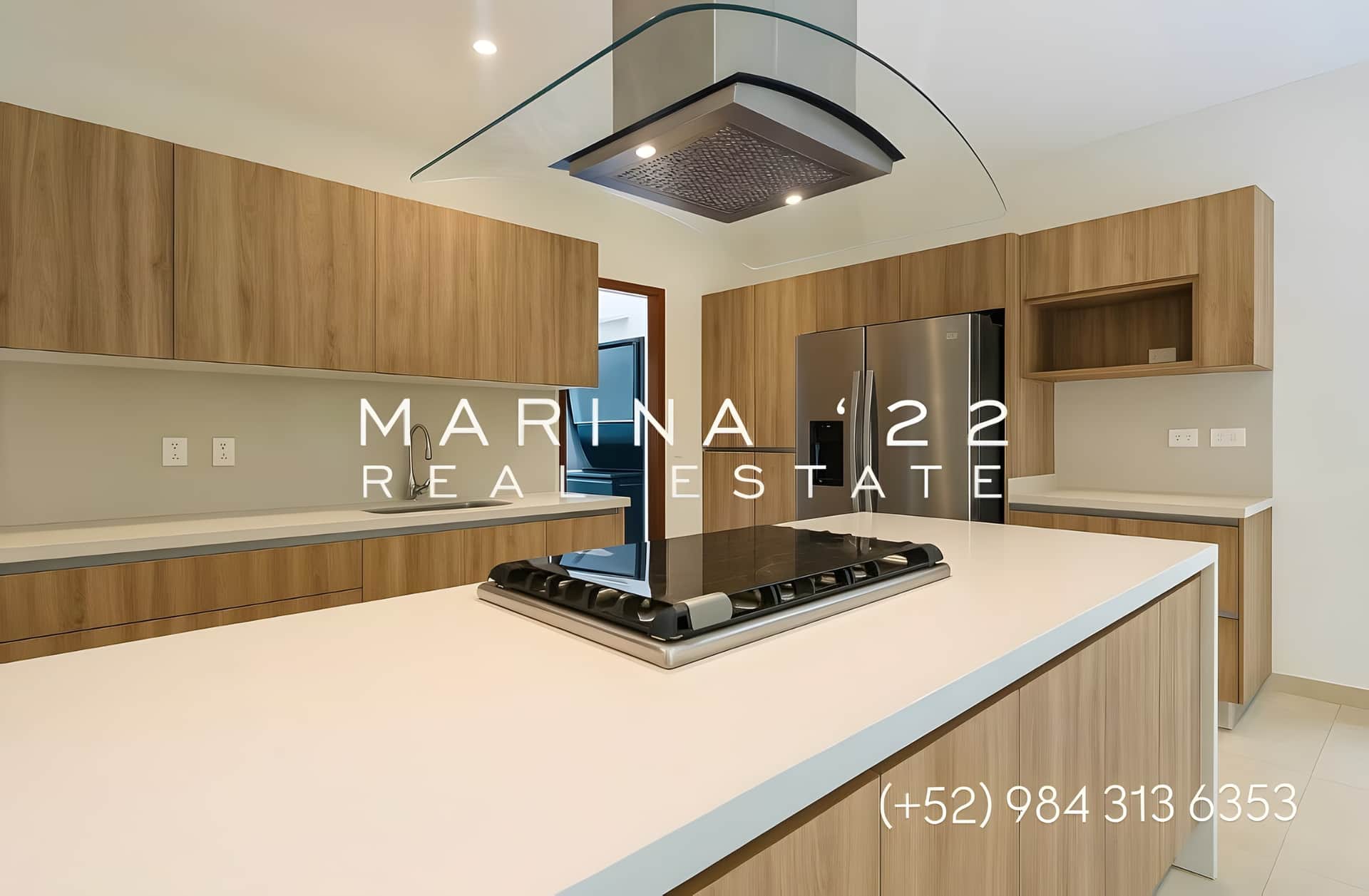 Cocina moderna con isla central, estufa empotrada, campana extractora de cristal, refrigerador de acero inoxidable y gabinetes de madera en tono claro.