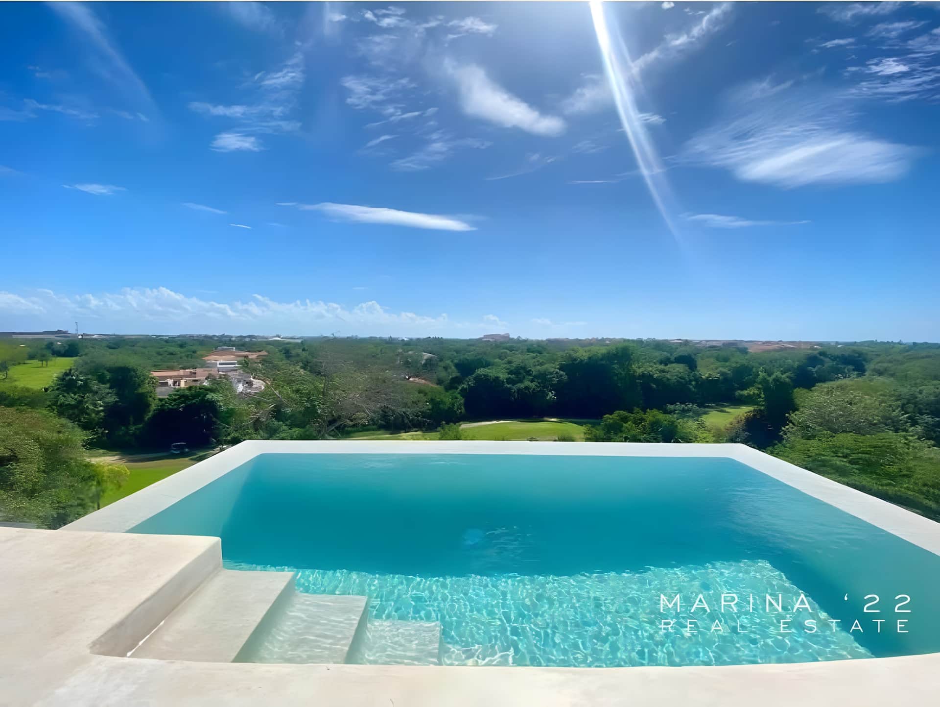 Alberca de borde infinito con escalones de acceso, vista hacia zona arbolada con algunas edificaciones y el mar al fondo bajo cielo azul con nubes dispersas.